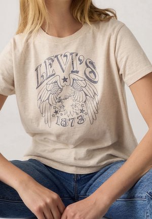 Kobieta w beżowym t-shircie Levi's z grafiką orła i tekstem "1873", w połączeniu z niebieskimi dżinsami, siedzi z rękami spoczywającymi na kolanach.