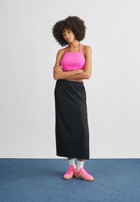 Rosa stickad halterneck crop top, svart maxikjol, rosa sneakers med vita detaljer och ankelsockor. Modellen står med armarna i kors mot en neutral bakgrund.