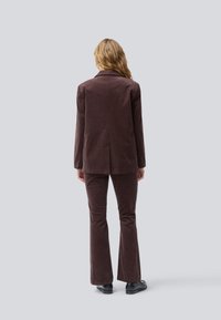 Donna con capelli biondi ondulati indossa un completo di velluto a coste marrone e scarpe nere, in piedi di spalle contro uno sfondo grigio chiaro semplice.