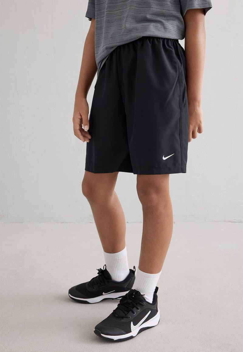 Schwarze Sportshorts mit elastischem Bund, kombiniert mit schwarzen Sportschuhen mit einem weißen Swoosh. Graues, gestreiftes Shirt und weiße Socken sichtbar.
