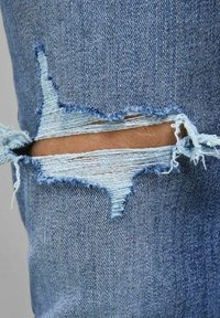 I jeans in denim blu presentano uno strappo grande e irregolare al ginocchio, che mette in evidenza un tessuto blu chiaro sfilacciato sottostante. I bordi usurati sono visibili.