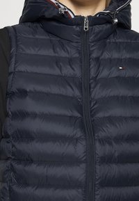 Granatowa pikowana kamizelka z zamkiem z przodu, poziomym przeszyciem, regulowanym kapturem i srebrnymi dodatkami. Na piersi znajduje się detal z logo.