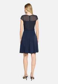 Robe marine avec un haut court, scintillant et transparent, et une jupe évasée. Se distingue par une ceinture et des plis ; associée à des sandales à talons hauts noires.