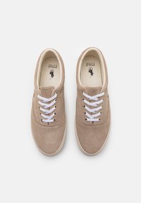 Beżowe zamszowe sneakersy z białymi sznurowadłami, okrągłym noskiem i teksturowaną gumową podeszwą. Logo nadrukowane na wkładce, klasyczny design z minimalnymi akcentami.