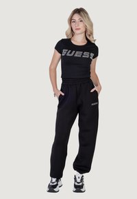 Must ülejäänud t-särk "GUESS" logo säravate kividega, koos lõdva musta dressipüksiga, millel on küljetaskud ja kitsenevad varrukad.