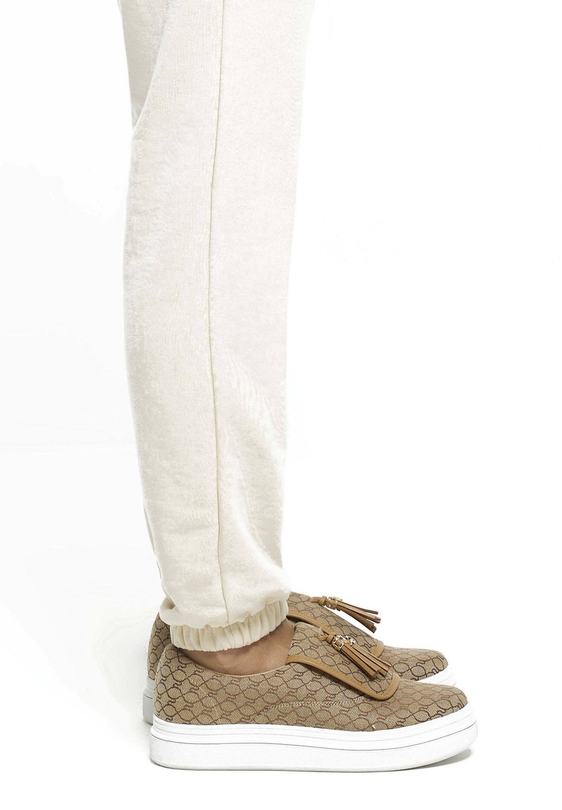 Personne portant un pantalon couleur crème et des baskets à enfiler marron à motifs avec semelles blanches et détails à pompons sur fond blanc.