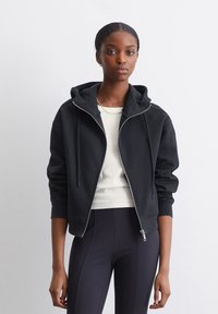 Sudadera negra con cremallera y una gran capucha, cierre de cordón y puños acanalados. Lleva puesta una blusa crema y pantalones negros ajustados. Textura suave.