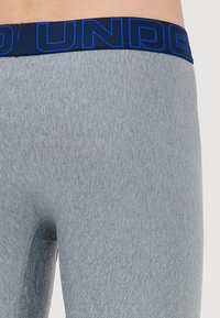 Primo piano di una persona che indossa un paio di pantaloni corti compressivi Under Armour grigio chiaro con una vita blu navy caratterizzata da lettere blu.