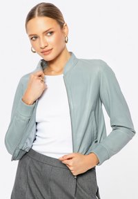 Lichtgrijze leren bomberjack met ribgebreide manchetten en kraag. Voorkant met rits en zijzakken. G gedragen over een witte top en grijze broek.