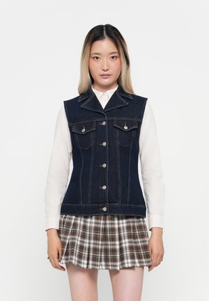 Donkerblauwe denimvest met een puntige kraag, drie knopen en vier voorzakken. Gecombineerd met een geruite geplooide rok.
