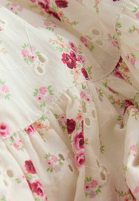Svetlo krémová látka so zoskupenými švami, vyšívanými dierkami a malými ružovými a červenými kvetinovými vzormi.