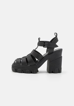 Sandalias negras con tacón bloque y múltiples correas anchas, hebilla en el tobillo y plataforma texturada con bordes dentados.