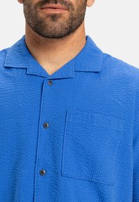 Camisa azul de botones texturizada con un bolsillo, que presenta un cuello tipo camp y botones negros visibles, destacando su patrón y diseño únicos.