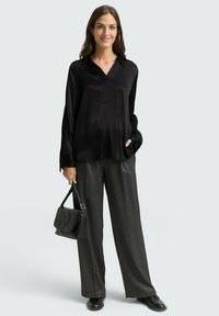 Blouse en satin noir avec un col, manches longues ; associée à un pantalon large gris scintillant taille haute ; tient un sac à main noir.