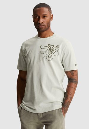 Homme avec des tatouages portant un t-shirt clair avec un imprimé floral abstrait et le texte « Cast Iron », debout les mains dans les poches.