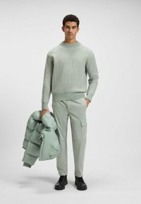 Lichtgroene gebreide trui met een ronde hals, gecombineerd met lichtgroene cargo-pantalons. Houdt een fluffy mintkleurige jack vast. Zwarte veterschoenen.