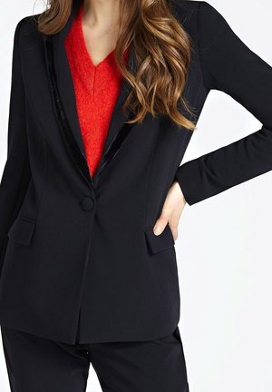 Kobieta w czarnym blezerze z welwetowymi klapami, pod którym ma jasnoczerwoną sweter z dekoltem w serek, ręka oparta na biodrze, widoczne długie falujące włosy.