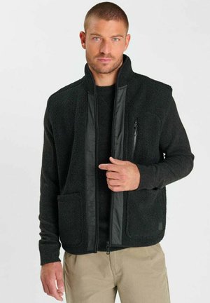 Uomo che indossa una giacca in pile nera sopra una camicia nera e pantaloni beige, in piedi davanti a uno sfondo chiaro uniforme.