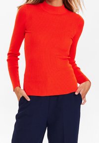 Gestreiftes Langarmoberteil in leuchtendem Orange. Verfügt über einen hohen Kragen und ein figurbetontes Design. Kombiniert mit dunklen Marinehosen. Weicher Stoff.