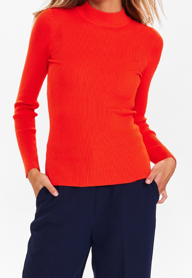 Gestreiftes Langarmoberteil in leuchtendem Orange. Verfügt über einen hohen Kragen und ein figurbetontes Design. Kombiniert mit dunklen Marinehosen. Weicher Stoff.