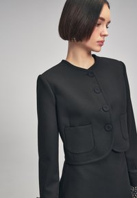 Blazer corto nero realizzato in tessuto liscio, con quattro bottoni, due tasche frontali e scollatura arrotondata con spalle sagomate.