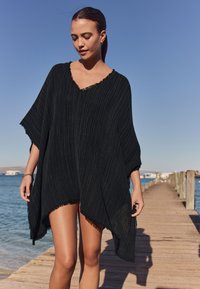 Črni pončo z resicami in V-izrezom, s teksturiranim črtastim vzorcem in ohlapnim krojem, nošen na modelu ob vodi.