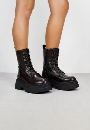 Botas de tobillo de cuero marrón con una suela de goma negra gruesa, parte frontal con cordones y cremallera lateral; con una lengüeta en la parte posterior.