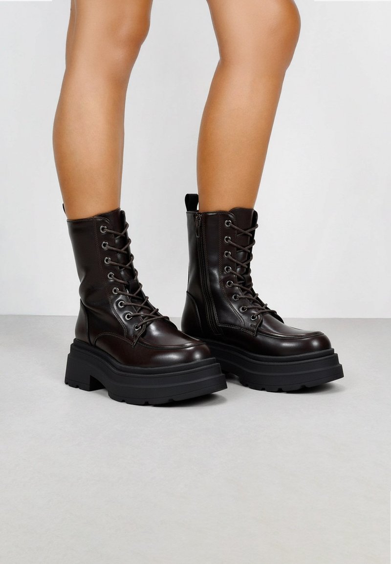 Botas de tobillo de cuero marrón con una suela de goma negra gruesa, parte frontal con cordones y cremallera lateral; con una lengüeta en la parte posterior.