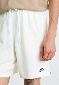 Balti medvilniniai šortai su juosmens raišteliu, su mažai juoda Nike logotipu ir šoninėmis skeltelėmis, derinami su bežine palaidine.