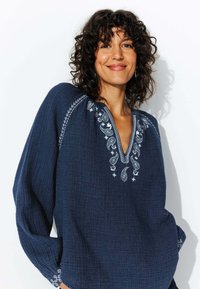 Blouse tissée bleu marine avec un décolleté en V, ornée de broderies en paisley blanches et de motifs floraux le long du col et des manches.