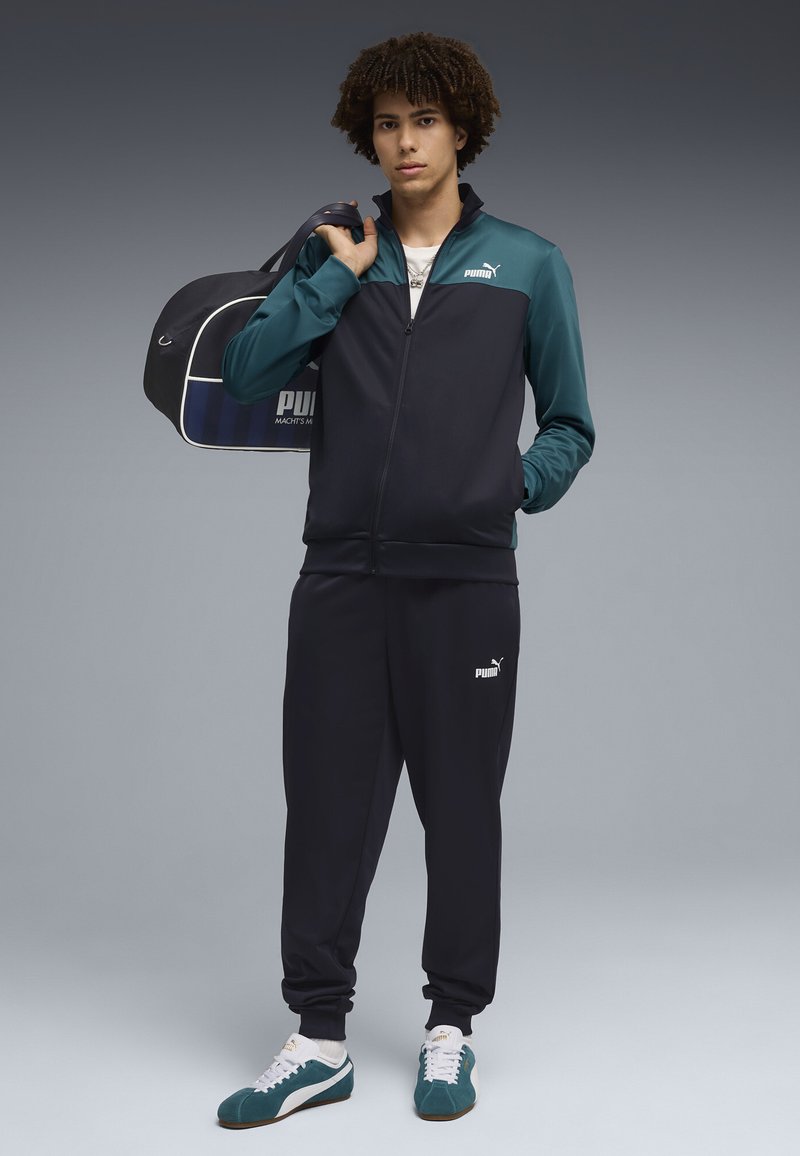 Jeune homme en survêtement Puma noir et vert, tenant un sac de sport Puma noir sur l'épaule, debout devant un fond gris.
