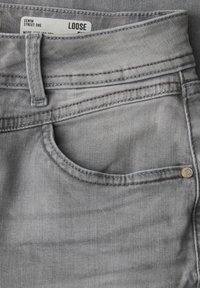 Bolsillo de jeans de mezclilla gris claro con presilla de cinturón visible y remache de metal, mostrando etiqueta blanca que dice "LOOSE" en la pretina.