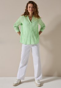 Camicia a righe verdi con colletto e maniche a tre quarti, abbinata a pantaloni bianchi a gamba larga e sneakers di colore chiaro.