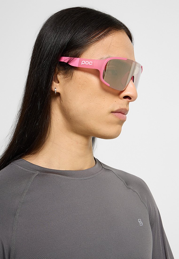 Różowe sportowe okulary przeciwsłoneczne z szeroką oprawą i lustrzanymi soczewkami, noszone przez osobę w szarej koszuli, z logiem marki na ramieniu.