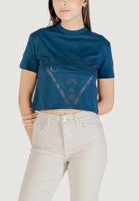 T-shirt corto color turchese con un logo triangolare nero lucido che include il nome del marchio "GUESS" e il testo "WASHED JEANS". Abbinato a pantaloni beige chiaro.