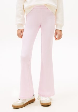 Dítě oblečené v světle růžových žebrovaných rozšířených kalhotách, bílém svetru a bílých teniskách s béžovými a hnědými podrážkami, stojící na bílém pozadí.