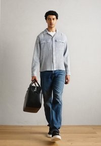 Blå og hvit stripete skjorte med knapper og to frontlommer, kombinert med blå jeans og svarte joggesko, mens han holder en svart veske.