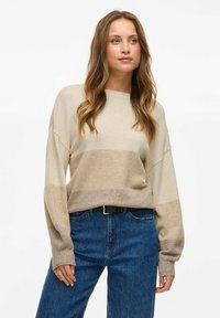Beżowy sweter dzianinowy z poziomym wzorem gradientowym, z okrągłym dekoltem i opadającymi szwami na ramionach, zestawiony z niebieskimi dżinsami.