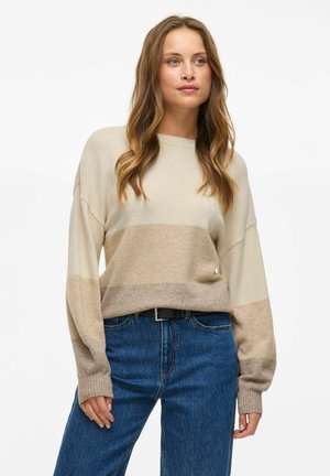 Pull en tricot beige avec un motif en dégradé horizontal, doté d'un col rond et de coutures d'épaule tombantes, associé à un jeans bleu.