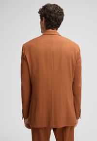 Ein rostfarbener Anzugjacke aus strukturiertem Stoff, mit scharfen Revers und einem einzelnen Schlitz. Sie verfügt über zwei Taschen und Knopfakzente an den Ärmeln.