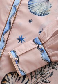 Manche de chemise avec poignet boutonné sur tissu rose clair présentant des motifs bleus de coquillages, étoiles, cordes et récifs coralliens.