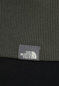 Tejido de rayas verde oscuro con una etiqueta cosida gris que muestra el logotipo de "THE NORTH FACE" y un ícono de montaña blanca en el borde inferior.