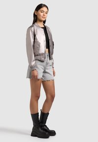 Jeune femme debout portant une veste bomber argentée brillante, un crop top noir, un short en denim clair et des bottes de combat noires à lacets.