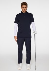 Tumman sininen polo-paita lyhyillä hihoilla, valkoinen pitkähihaisinen paita alla, yhteensopivat tummansiniset housut, valkoiset lenkkarit ja golfmaila kädessä.