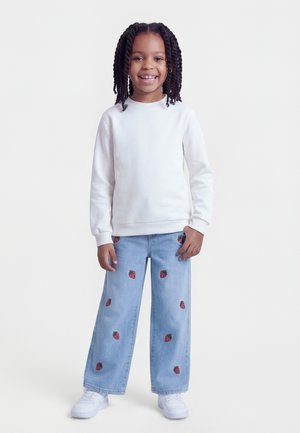 Mladé dievča s vrkočmi, oblečené v bielej tričku s dlhým rukávom, svetlomodrých džínsach s potlačou jahôd a bielych teniskách, stojace a usmievajúce sa.