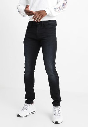 Osoba nosząca białe trampki, wąskie czarne jeansy oraz białą koszulę z długim rękawem z nadrukami na rękawach.