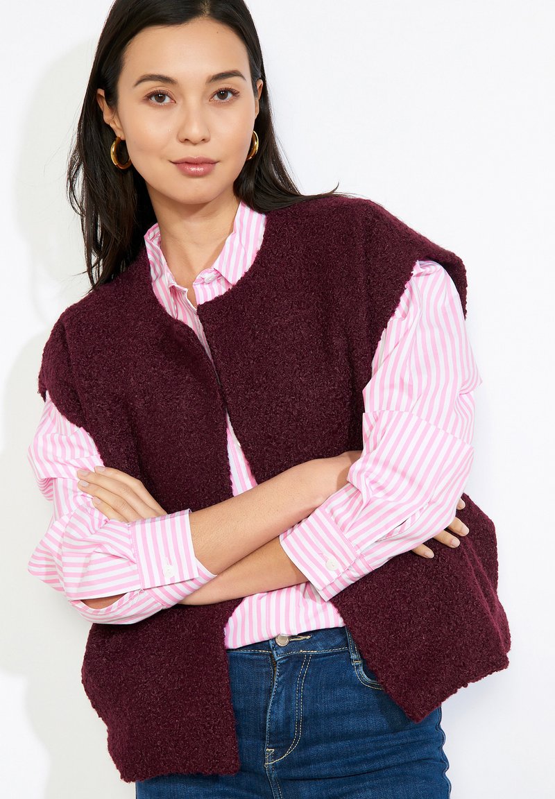 Mujer con pendientes de aro dorados, camisa a rayas rosas y blancas, chaleco sin mangas texturizado color burdeos y vaqueros azules con los brazos cruzados.