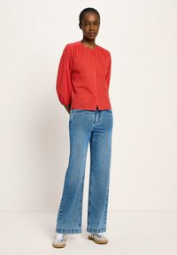 Frau steht vor einer beigen Wand, trägt eine rote Knopfbussinessbluse, weit geschnittene blaue Jeans und weiße Sneakers, mit den Händen hinter dem Rücken.