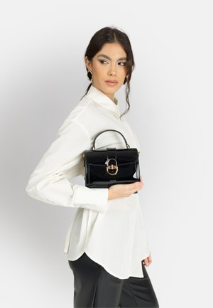 Donna con camicetta bianca e pantaloni neri che tiene una borsa nera lucida con chiusura dorata, guardando di lato su uno sfondo semplice.