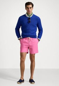 Άνδρας που φοράει μπλε πλεκτό πουλόβερ, λευκό πουκάμισο, πράσινο μαντίλι λαιμού με σχέδια, ροζ σορτσάκι με κορδόνια, ναυτικές εσπαντρίγιες και γυαλιά ηλίου κρεμασμένα στο πουλόβερ.